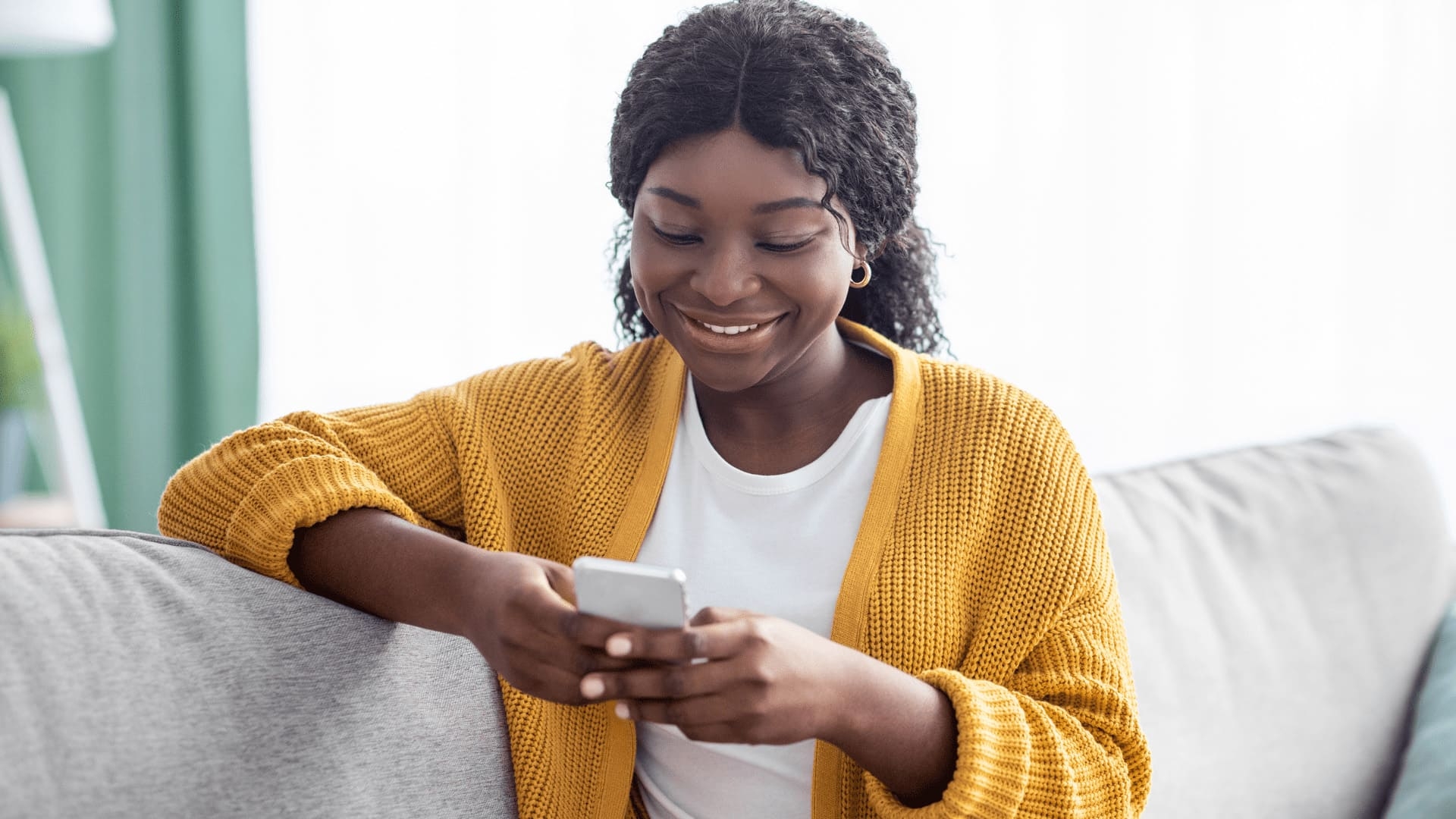 Smiling woman in a yellow sweater sitting on a couch, using her smartphone