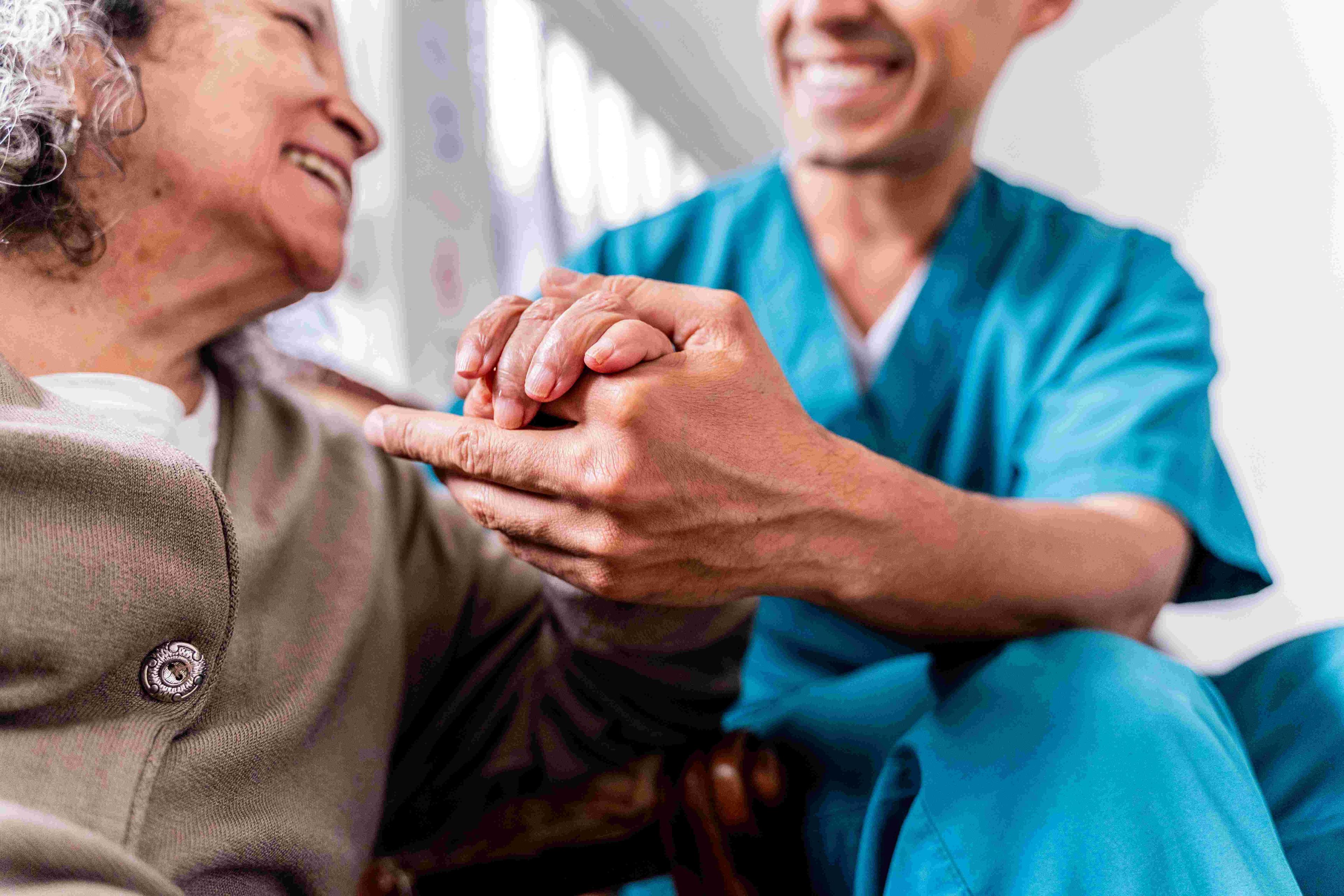 A close-up shot of a male caregiver in blue scrubs sitting with and holding the hands of a smiling senior woman