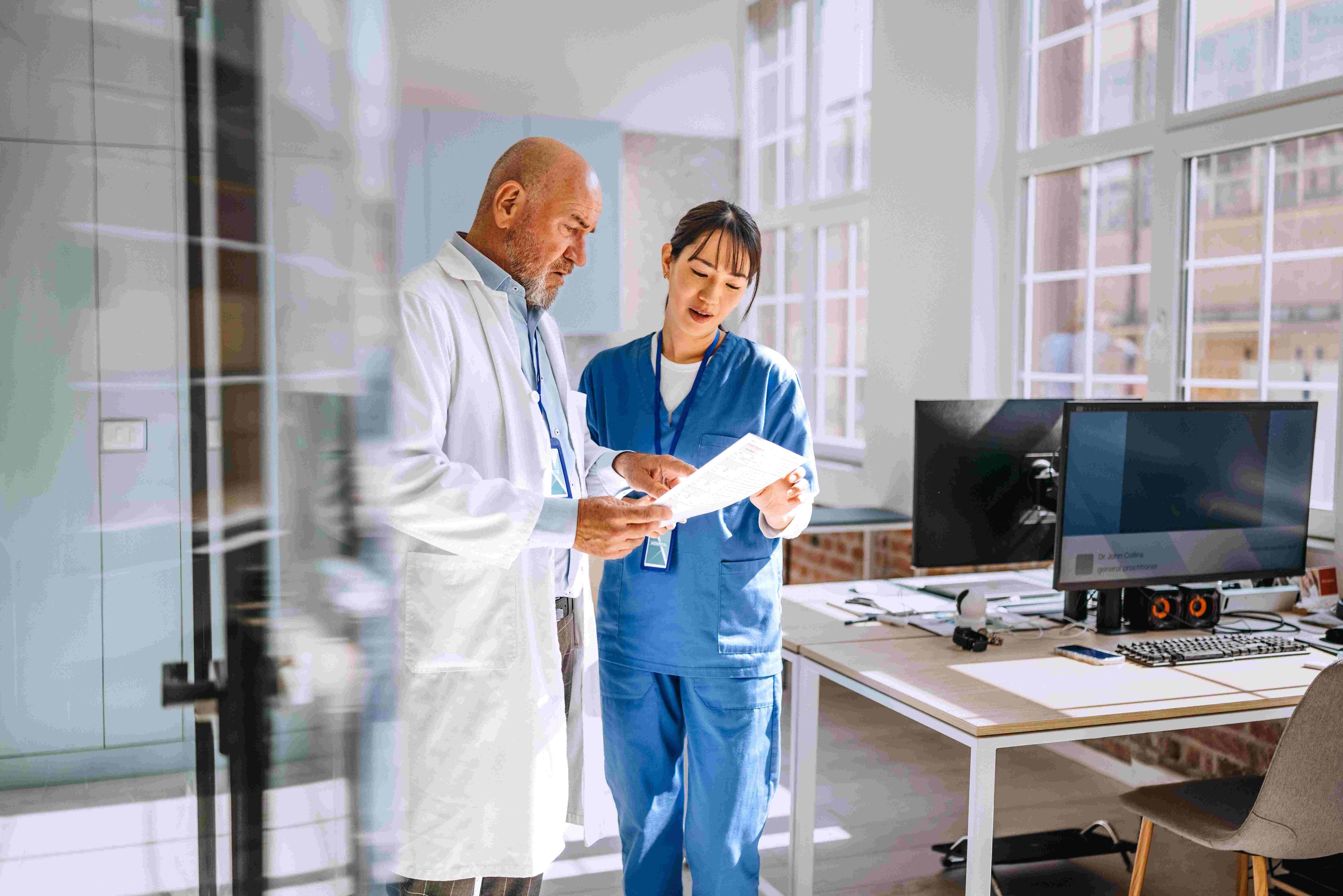 An experienced male doctor in a white lab coat and a younger female medical professional in blue scrubs are standing and reviewing a document together in an office.