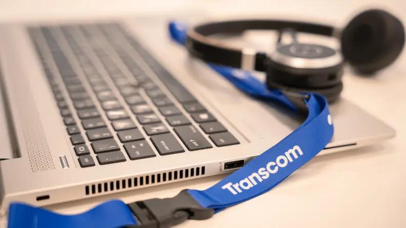 A close-up of a laptop keyboard with a blue 'Transcom' lanyard and a black headset resting on top.