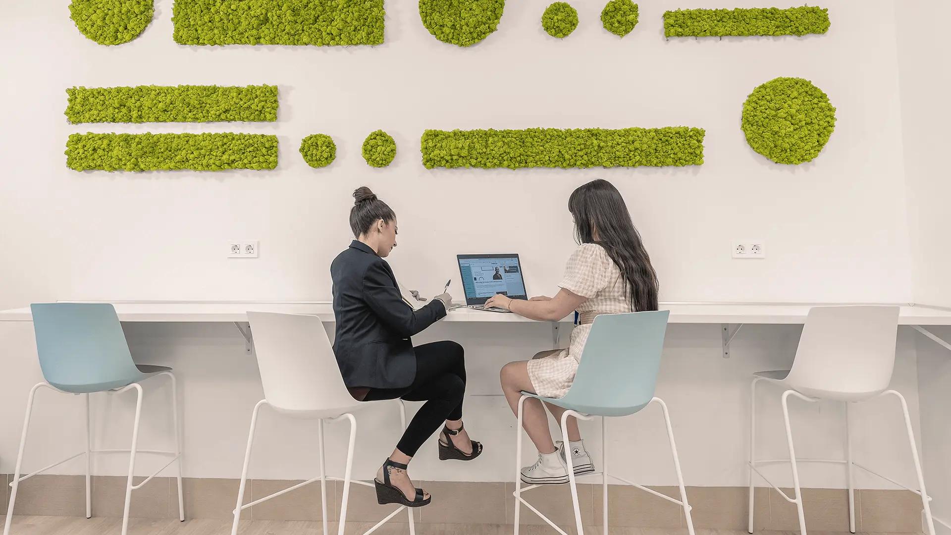 Two women sitting in a modern workspace, collaborating on a laptop with a minimalist white desk and green wall decor in the background.