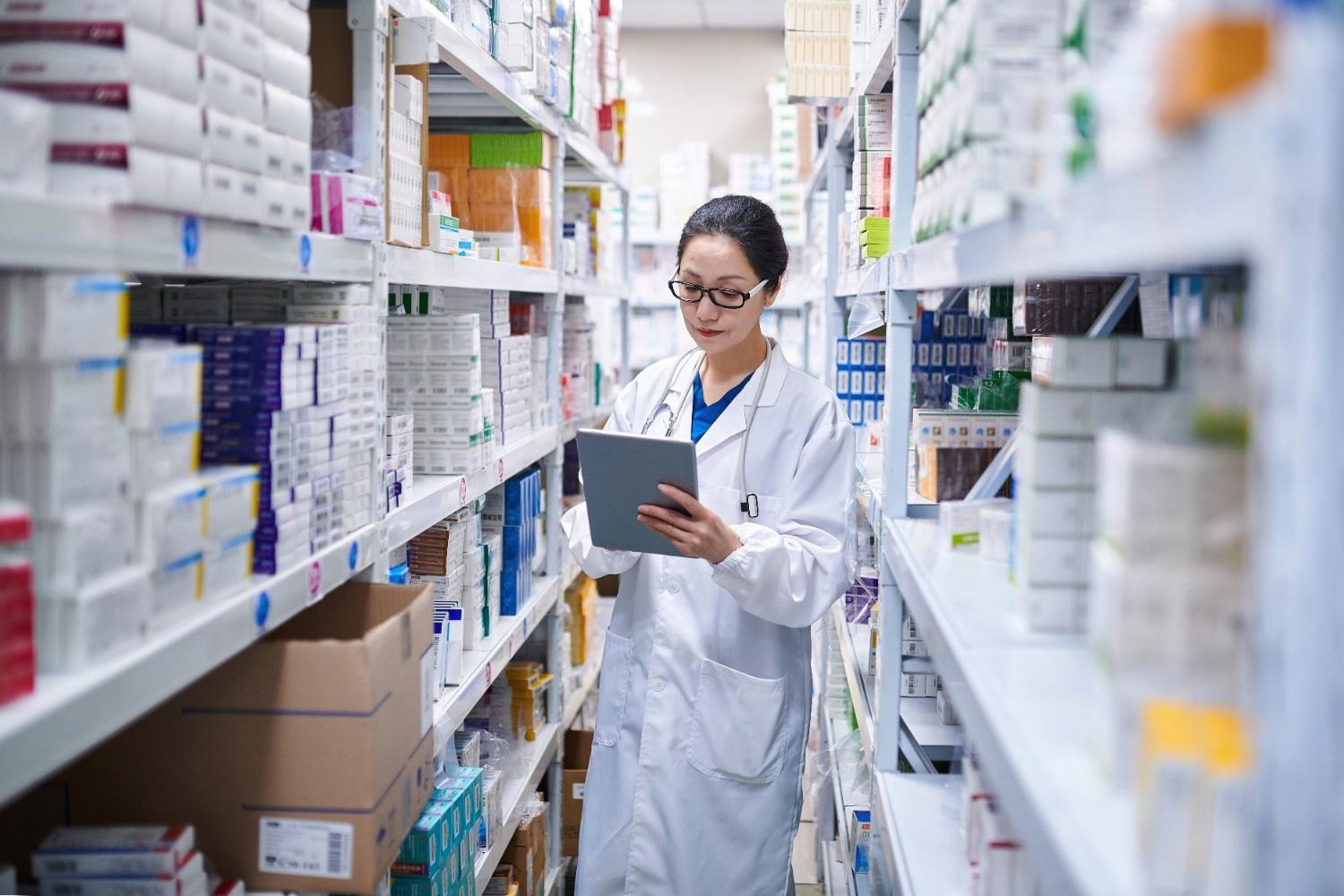A female pharmacist in a white lab coat is standing in an aisle of a pharmacy, using a tablet to check inventory or manage stock on the shelves.