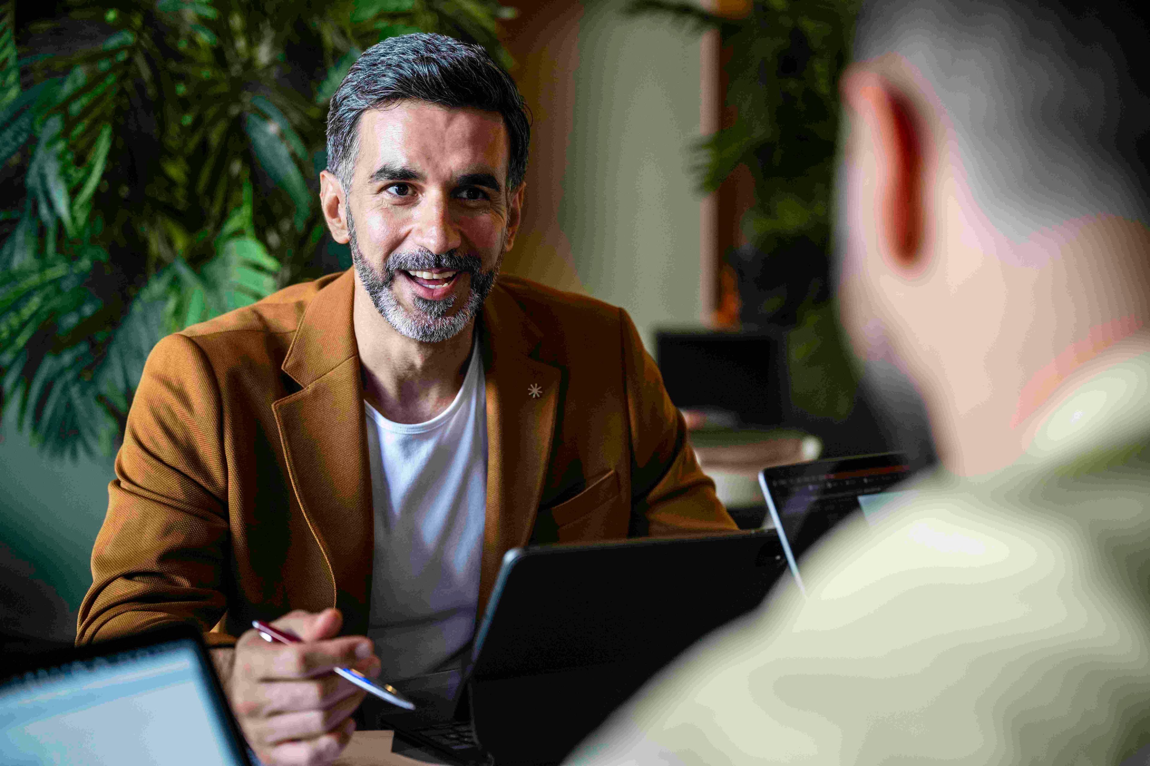 A close-up shot of a smiling man with a beard and brown jacket sitting at a table with a laptop, looking at someone off-camera during a business meeting.