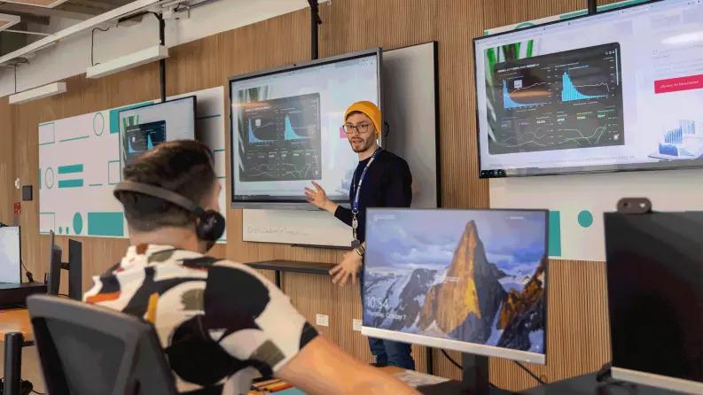 A presenter explaining data on multiple screens to an audience in a tech-focused training session