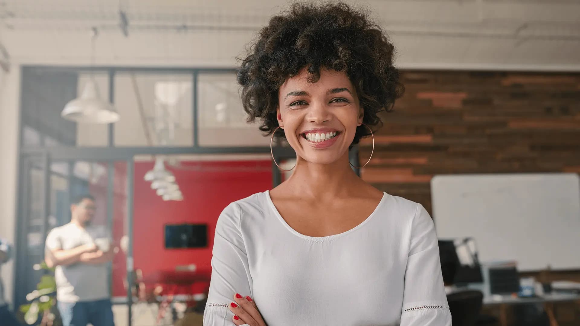 A smiling woman with curly hair, wearing a white blouse, standing confidently in a modern office space with colleagues in the background.