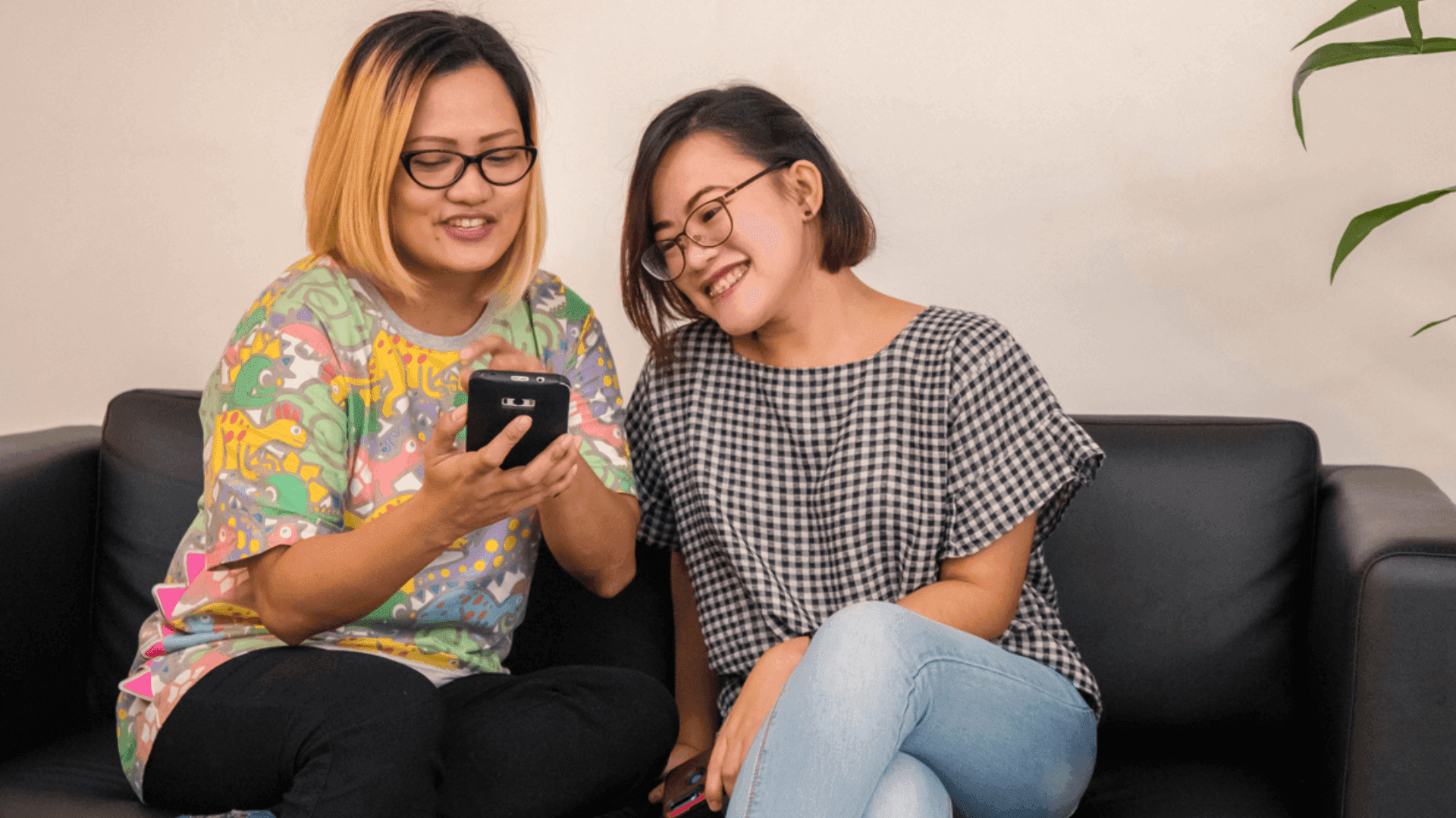 Two women smile as they sit together and use a mobile device.