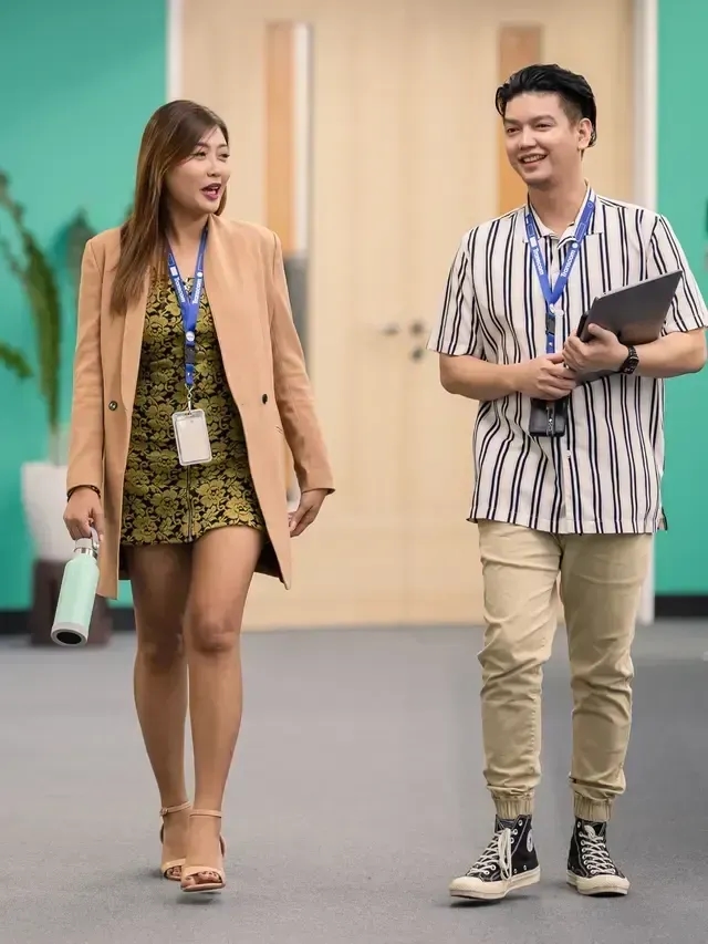 Two professionally dressed colleagues walking and talking in an office hallway, both wearing ID badges.